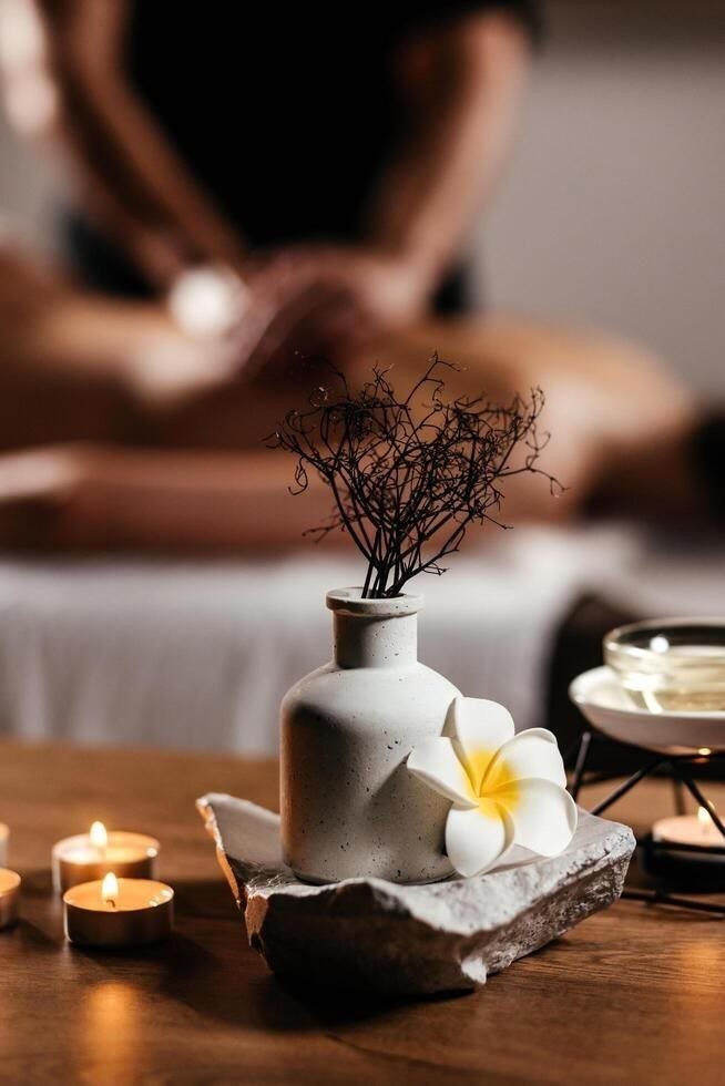 Candlelit spa table with a ceramic vase, frangipani flower and massage oil, with a massage session in progress in the background at Blossoms Massage Spa